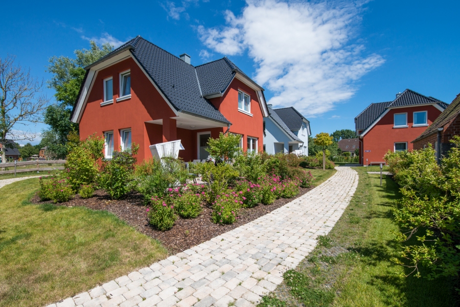 Ferienhaussiedlung mit drei Holzhäusern in Schwedenfarben – Rot und Blau – mit weißen Fensterrahmen, Terrassen und gemütlichen Strandkörben unter blauem Himmel.