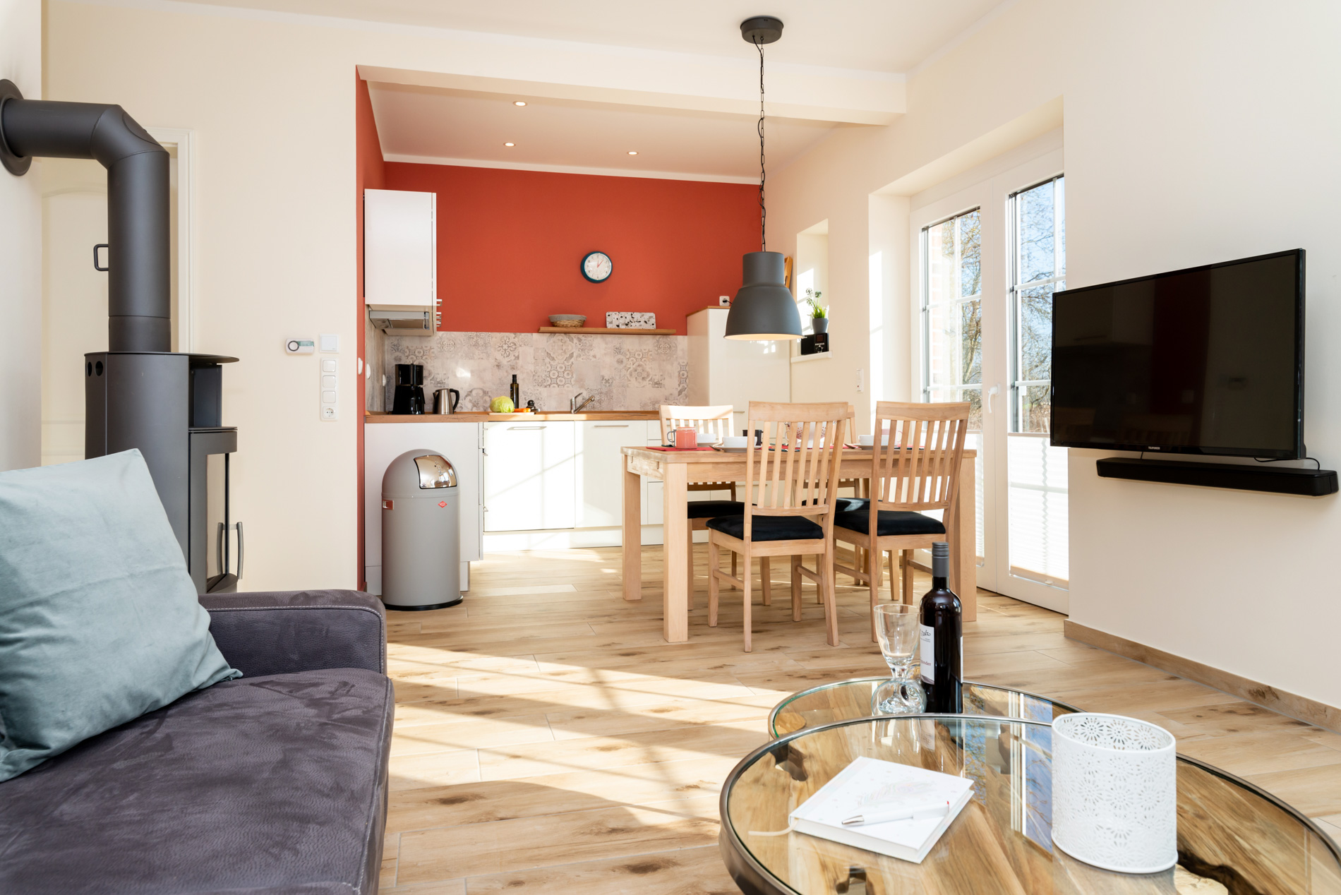 Modernes Wohnzimmer im Ferienhaus auf Fehmarn mit knisterndem Kaminfeuer, bereitgelegtem Kaminholz und hochwertiger Einrichtung auf hellen Bodenfliesen.