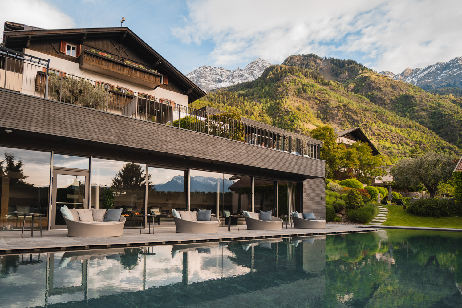 Außenansicht des Hotel Mair am Ort Living in Partschins bei Meran in Südtirol mit großzügigem Außenpool, stilvollen Loungebereichen und gepflegtem Garten vor der beeindruckenden Bergkulisse des Naturparks Texelgruppe. _fullwidth-story-splash