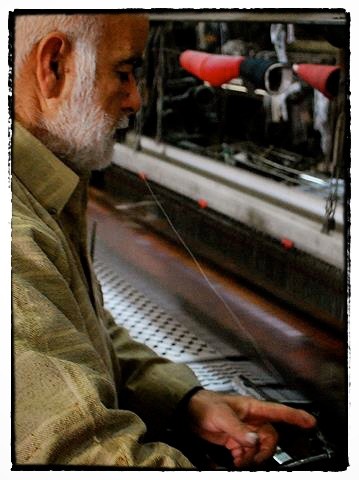 While a loom pounds out a traditional black-and-white kufiya, Abid Keraki holds a thread in his mouth as he quickly replaces a spool. The eight operational looms require constant attention.