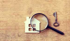 A magnifying glass looking at a house on a wooden background with a key.