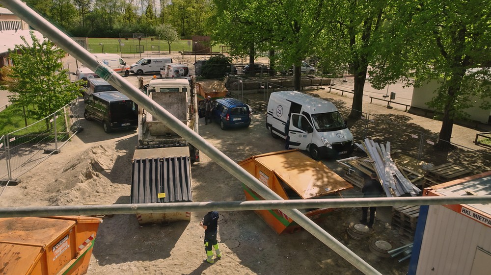 Hier sieht man, wie viele Leute auf der Baustellearbeiten. Haufenweise Autos auf der Baustelle. Auf der Straße und den Parkplätzen stehen auch noch welche.