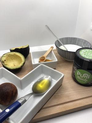 Photo de plusieurs ingrédients, huile d'olive, avocat, yaourt blanc, miel de manuka, pour fabriquer sa propre crème hydratante au miel de manuka sur une planche en bois sous fond blanc