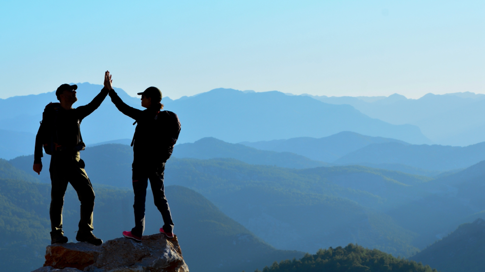 Zwei Menschen, die mit erhobenen Armen sich abklatschen auf einem Berggipfel, symbolisiert Selbstermächtigung, die Gemeinschaft und Erfüllung des inneren Potentials.