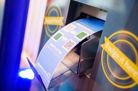 Nahaufnahme des Ausgabeschachts eines SelfPrint-Kiosks. In dem Ausgabeschacht liegt ein fertig gedrucktes Badge.