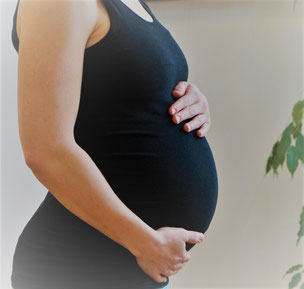 Das Bild zeigt eine Schwangere mit Babybauch in der Seitenansicht. Die Hände sind liebevoll auf den Bauch gelegt. Im Hintergrund befindet sich eine Pflanze.