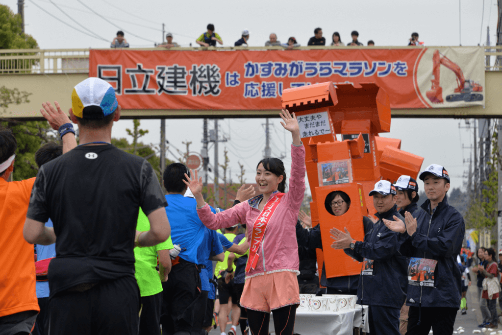 【第25回大会（2015年）】ランナーを応援するミス日本とメインスポンサーである日立建機のボランティアの皆さん。 重機をイメージした被り物も、今では「かすみがうらマラソン」きっての名物になりました。
