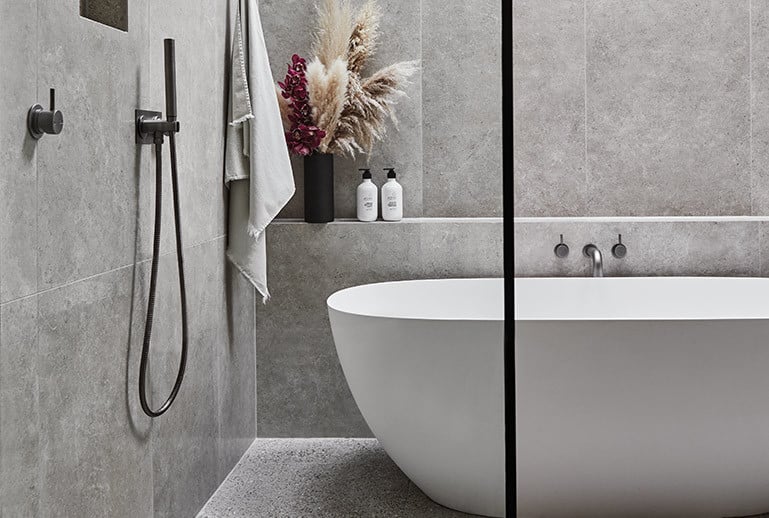 A serene bathroom features a large white freestanding bathtub with sleek gunmetal mixers, a skylight that frames the sky, and expansive grey tiles with matching grout, evoking a tranquil forest ambiance.