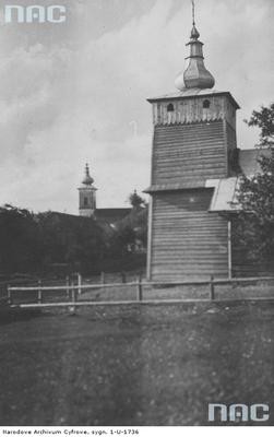 Izby - prawosławna cerkiew drewniana, w tle widoczna wieża cerkwi św. Łukasza. Zdjęcie pochodzi ze zbiorów Narodowego Archiwum Cyfrowego