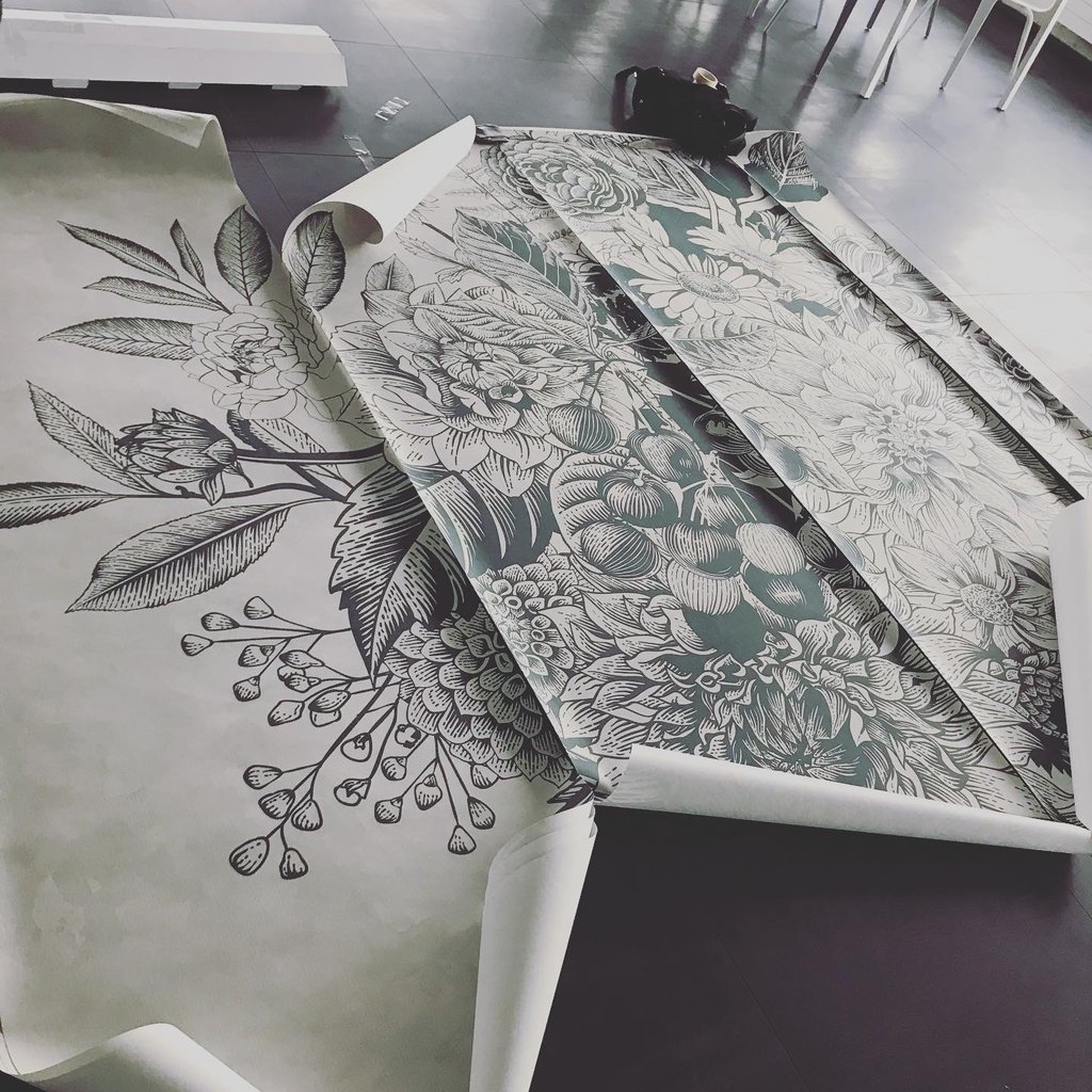 Tapetenbahnen liegen auf dem Boden Ausgebreitet. Grafisch umgesetztes Pflanzen-Muster mit Blättern, Blüten und Früchten in Grün auf Beige.