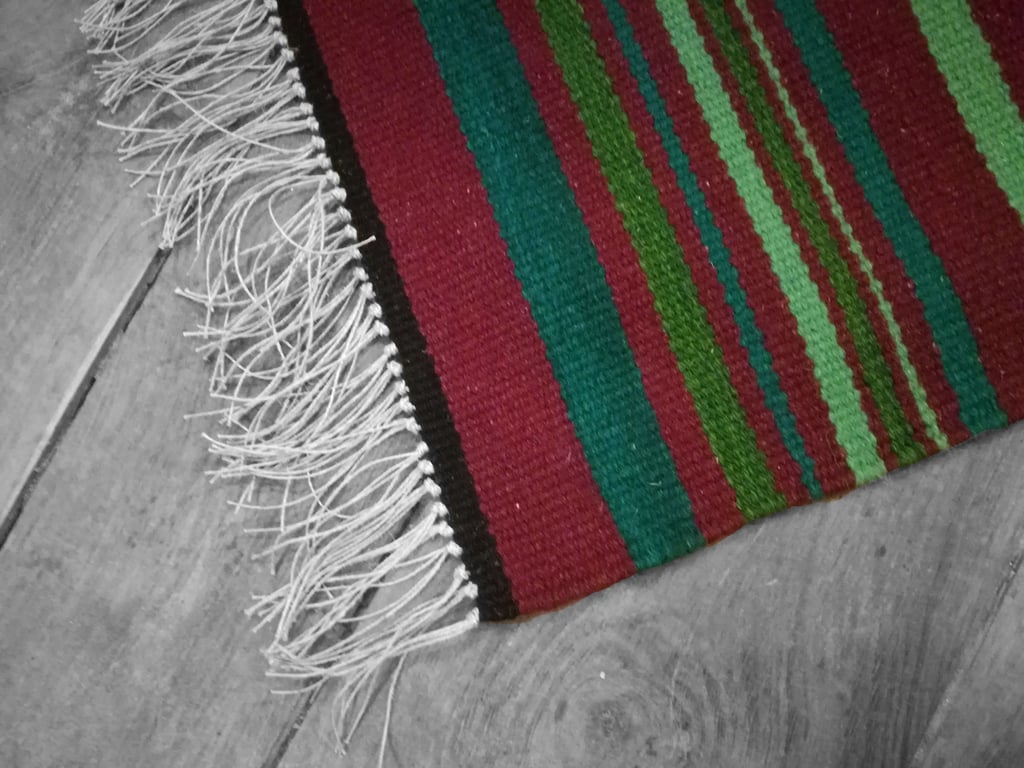 On voit un détail du tapis - descend de lit - rouge bordeaux aux rayures irrégulières vertes est posé sur le parquet en bois. Les franges en fils de lin son en premier plan 