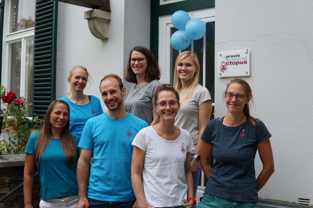 Das Team des Physioterapiezentrums Praxis Octopus steht vor dem Eingang zu den Räumlichkeiten. Alle Therapeutinnen lachen freundlich und strecken die Arme in die Luft.