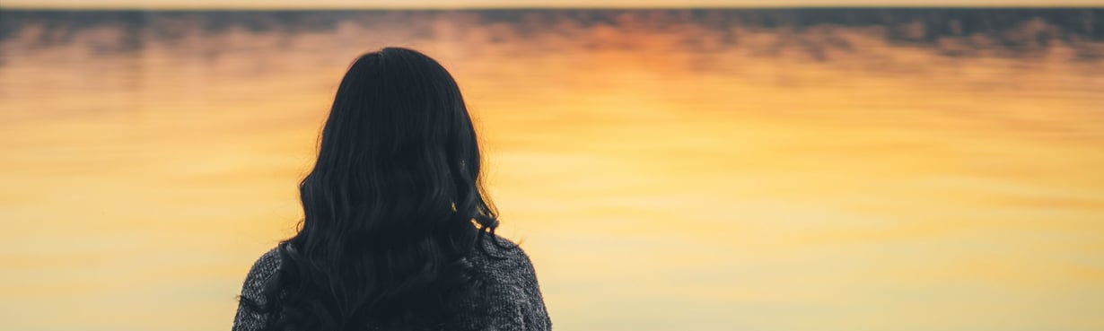 Frau sitzt mit dem Rücken zur Kamera an einem ruhigen Gewässer im warmen Abendlicht, ein Moment des Innehaltens und inneren Übergangs.