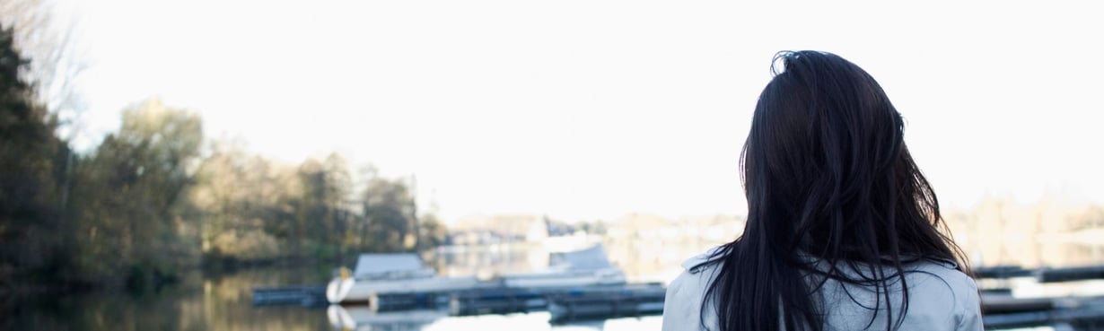 Frau von hinten steht still an einem ruhigen Gewässer, Blick in die Weite als Symbol für einen inneren Übergang und einen Raum, der Halt gibt.