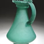 Einhenkelkanne aus blau-grünem Glas mit einem geripptem Bandhenkel und auf dem Mundrand verschmolzenen, gekniffenen Glasfäden.  1. Hälfte 4. Jh.n.Chr.  Fundort: unbekannt  Inv. Nr. G 678 a  Höhe ca 165 mm  Gefäß Nr. 706