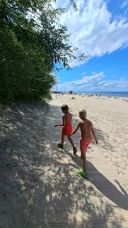 Two boys in orange swimming trunks walking along tree-fringed beach with blue sky in Jūrmala, Latvia