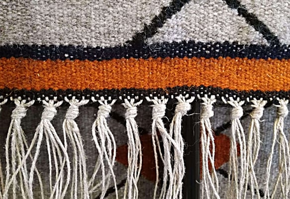 Détail de la lisière d'un grand tapis kilim tissé à la main en laine, au fond gris clair, avec un motif losange en noir et orange. Au bord du tapis, les fils de la chaîne sont noués en frange.