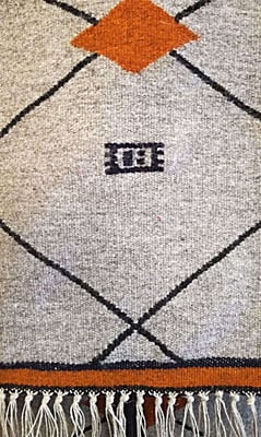 Détail de la lisière d'un grand tapis kilim tissé à la main en laine, au fond gris clair, avec un motif losange en noir et orange. Une signature - logo de tisserand est en centre. 