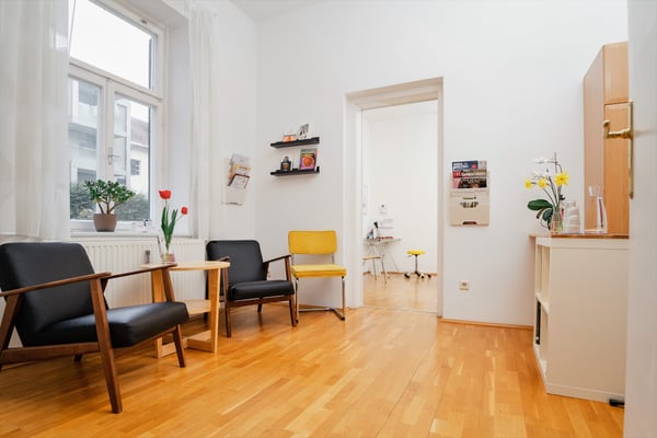 Das Bild zeigt einen hellen Warteraum mit gelben und schwarzen Sesseln. Im Fenster steht eine Pflanze und es sind Blumen aufgestellt. Der Raum lädt zum Verweilen ein.