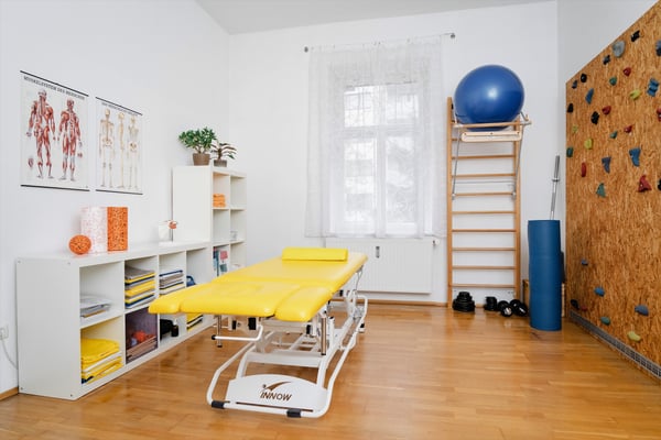 Ein gut ausgestatteter Therapieraum mit Sprossenwand, einer gelben Therapieliege, Gymnastikball, Yogamatte, Gewichten und Kletterwand. An der Wand hängen anatomische Bilder zur Aufklärung.