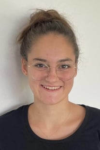 Portraitfoto von Sarah Schneck, Physiotherapeutin mit Schwerpunkt Kiefergelenk (CRAFTA). Sie hat eine große, runde Brille und die Haare am Kopf zusammengebunden. Sie trägt ein schwarzes Shirt, der Hintergrund ist grau.