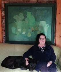 Painter Laila Kelle and her black dog in front of a green toned still life painting