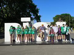 Group of teenage tourists in green shirts and guide with orange cap standing in front of white Riga sign