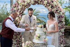 Bride and groom cutting wedding cake under a floral arch with ocean view in the background