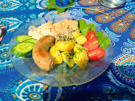 Clear glass plate with a sausage, boiled potatoes, tomatoes, lettuce and cucumbers on a blue tablecloth