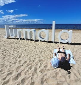 Man in black swimmers lying on back on beach in front of sign saying Jūrmala, Latvia