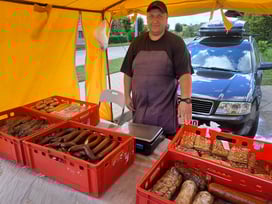 Man wearing a grey apron in front of red trays filled with homemade sausages in Dagda, Latvia