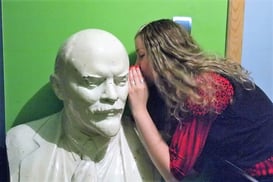 Latvian girl in red dress whispering into the ear of a bust of Lenin