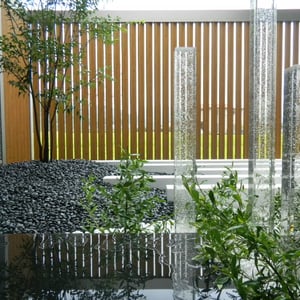 玄関を入ると正面に窓があり、坪庭が見えます。アルミ材のパーティション（正面奥）、ガラスの角柱、山西黒という天然石磨き材等を使い、現代和風デザインの坪庭になっています。天然石（手前下）は本磨き仕上げで日中は空のブルーや雲が映りこんだり、雨が降ると薄く水が張ったりしていろんな表情を楽しませてくれます。夜になるとライトアップされ、昼とは違う表情を見せてくれます。　　岡山市北区Y様邸　ガーデン　フラワーチルドレン