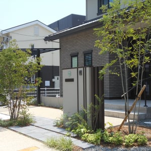 奥様がお気に入りの門壁イメージを元に、カスタマイズしました。重厚な佇まいの家に負けないよう、空に伸びる縦ラインを強調し、表札は後々取り替えられるユニソンのワンロックサインを採用しました。両サイドに植えたヤマボウシとシマトネリコが、自然の門構えとなってくれます。　　　　　　　　岡山市北区　H様邸　フラワーチルドレン　エクステリア