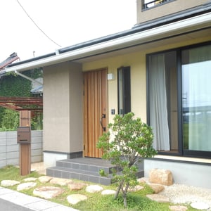 ナチュラルな感じで家の外観に合うものをというご希望に沿い、地元の山で採れる自然石を使って、玄関へのアプローチを作りました。また、玄関ドアの色合いに合わせた機能門柱を選び、真鍮製の表札一文字一文字はオリジナルで、スリット状門柱の躯体部分めがけてボルトを出し、裏からナットで締めています。リビング前には、ヤマモモに似たコロンとかわいい実が食べられるヒメイチゴノキを植えました。　　　　倉敷市　F様邸　フラワーチルドレン　エクステリア　　　　　　　　