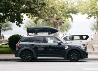 alt="Dunkles Fahrzeug mit Dachträger im Vordergrund, Bäume sind im Hintergrund zu sehen."