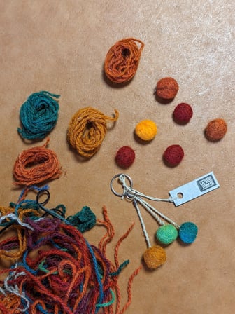 Plan de travail avec les fils de la laine de toute couleur mélangés, les balles en laine feutrée et un porte-clé avec trois petites pelotes de feutre - bleue, verte et marronne. 