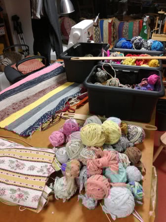 Préparation de la lirette - sur la table les pelotes de bandes de tissus découpés, le tapis en lirette, le tissu à découper, derrière les paniers noirs remplis de plus de pelotes. 