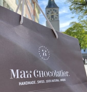 Im Vordergrund eine Tasche mit der Aufschrift Max Chocolatier. Im Hintergrund die Kirche St. Peter in Zürich. 