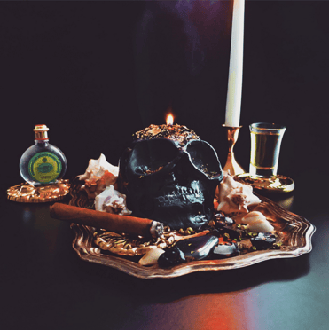 Eine zauberhafte schwarzer Schädel kerze ruht auf einem verzierten Teller, umgeben von Kerzen, Kräutern und einer kleinen Flasche 4711 eau de cologne