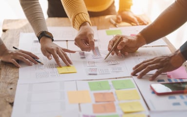 Mehrere Hände arbeiten gemeinsam an Strategien und Layouts auf einem Tisch – visuell als Symbol für Veränderungsprozesse in KMUs und Teams im Wandel.