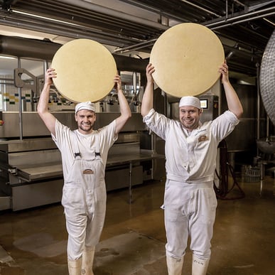 Zwei Personen in Hygienekleidung stehen im Produktionsbereich und halten Käse in den Händen