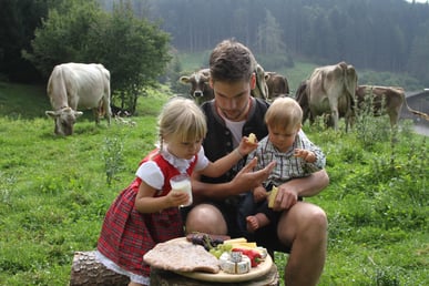 Ein Mann und zwei Kinder genießen Käse auf einer Almwiese, während Kühe im Hintergrund grasen.