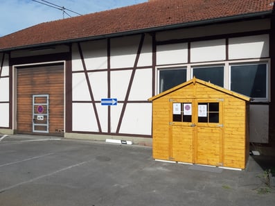 Rote kleine Holzhütte ohne Fenster, rechts daneben größeres Gebäude, links niedrige Hecke ohne Grün, davor geteerter Hof, im Hintergrund größeres Wohnhaus