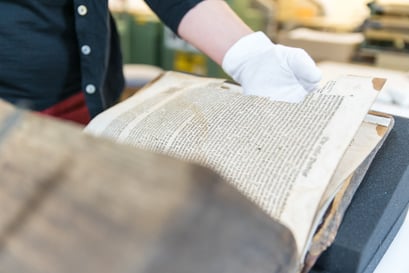 Katharina Lässig hält eine aufgeschlagene Bibel. Sie trägt zum Schutz des Objekts weiße Handschuhe.Die Bibel liegt auf Schaumstoffkeilen.