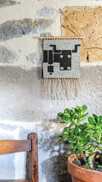 Sur une terrasse le kilim-tenture murale en tissage pixelisé représentant un écureuil sur le fond de la laine gris foncé est accroché sur un volet à côté de la fenêtre. Dessin du tissage mural géométrisé.