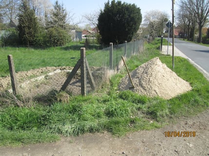 Begrenzungszaun, dahinter das Erdloch und daneben ein Sandhaufen mit Schaufel