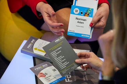 Eine Person präsentiert einer anderen ein Namensschild. Sie sitzen an einem Tisch.