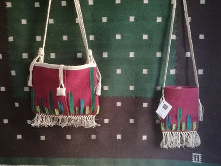 Deux sacs tissés en laine avec franges, couleurs bordeaux avec éléments verts , grande bandoulière et petit cabas, sont exposés sur un tapis kilim brun-vert avec les petits carrés gris clair.