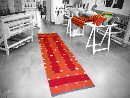 L'intérieur d'atelier de tissage la Trame avec un long tapis kilim au sol et d'autres tapis et coussins en exposition. Deux métiers à tisser de deux côtés sont visibles. Kilim est tissé à la main, il a fond rouge et orange et le motif des diamants gris.
